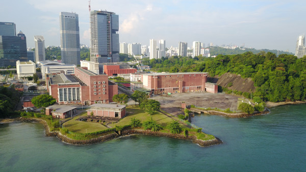 Aerial view of a red brick building complex on a coast, backed by skyscrapers and trees.